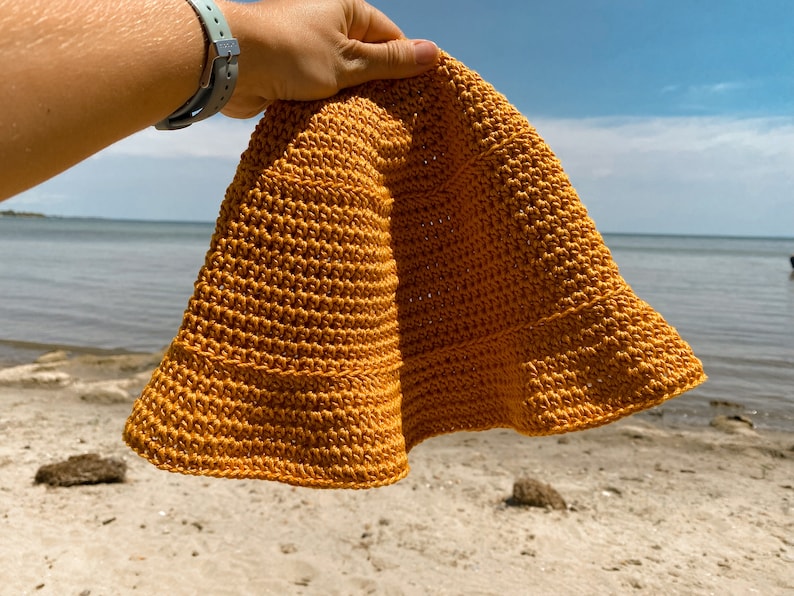 Op de afbeelding: Een gebreide bucket hat in een mosterdgele kleur. De hoed wordt in een hand gehouden tegen een achtergrond van een strand en blauwe lucht.