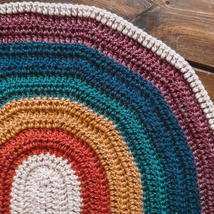 May include: A crocheted rainbow rug with a white center. The rug is made of different colored yarns, including red, orange, yellow, green, blue, and purple.