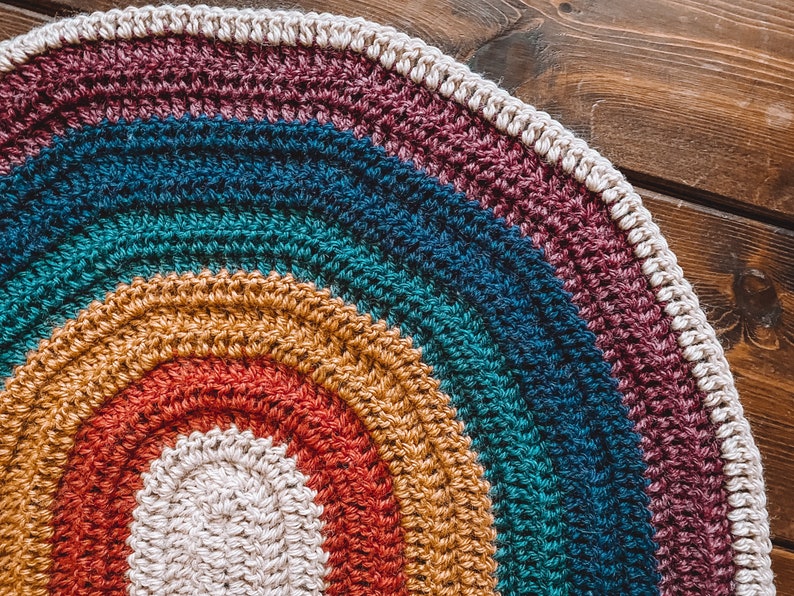 May include: A crocheted rainbow rug with a white center. The rug is made of different colored yarns, including red, orange, yellow, green, blue, and purple.