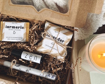 May include: A gift box filled with bath and body products, including a turmeric oatmeal scrub bar, a lavender bath bar, a small bottle of essential oil labeled "zen", and a glass tube of bath salts. The box is lined with brown paper shreds.
