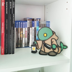 Puede incluir: Una figurita marrón y verde de un personaje que sostiene una linterna. La figurita está en un estante blanco con cajas de videojuegos detrás.