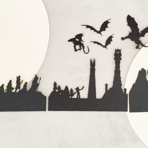 May include: Black silhouette paper cutouts of characters from the Lord of the Rings, including the Fellowship, the tower of Barad-dûr, and a dragon flying overhead.