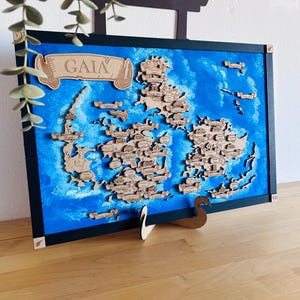 Può includere: Mappa in legno con sfondo dipinto di blu e bianco. La mappa presenta ritagli in legno di masse terrestri ed etichette. La parola "GAIA" è visualizzata su uno striscione. La mappa è incorniciata in nero e supportata da un supporto in legno.