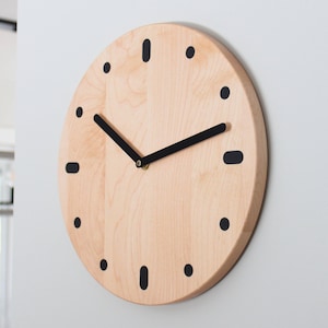 May include: A round wooden clock with black hour markers and black hands. The clock face is made of light wood with a natural grain pattern.
