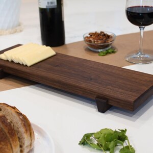 May include: A dark brown wooden serving tray with a raised edge and four small feet. The tray is made of walnut wood and has a natural finish. There are slices of cheese on the tray.