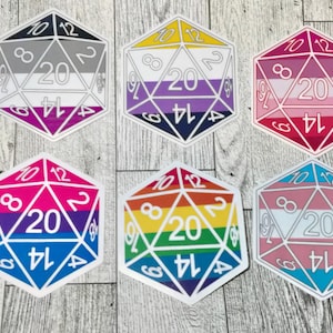 May include: Six colorful stickers in the shape of a twenty-sided die. Each die features a different LGBTQ+ pride flag design, with numbers and edges in white. The dice are set against a light wood grain background.