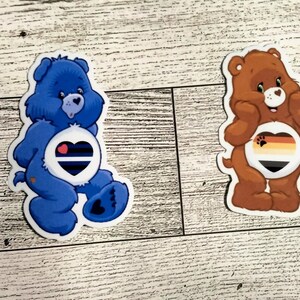 May include: Two Care Bear stickers with pride flags on their bellies. The blue bear has a blue, black, and white pride flag on its belly. The brown bear has a brown, orange, black, and yellow pride flag on its belly.