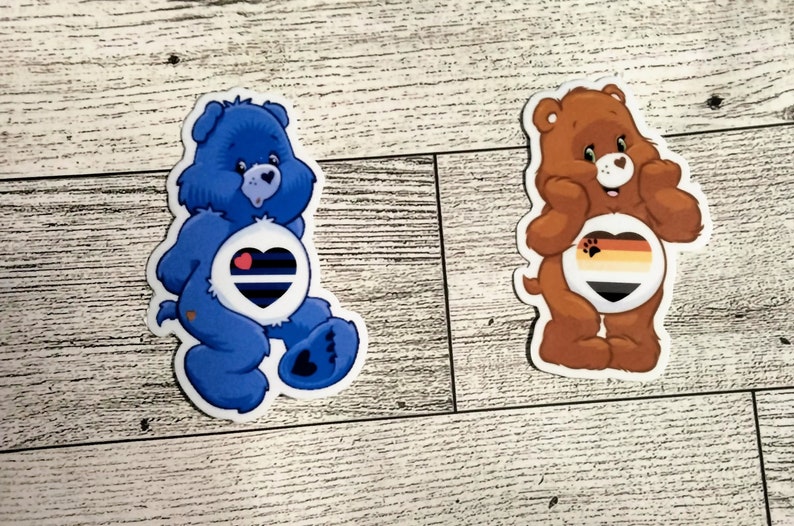 May include: Two Care Bear stickers with pride flags on their bellies. The blue bear has a blue, black, and white pride flag on its belly. The brown bear has a brown, orange, black, and yellow pride flag on its belly.
