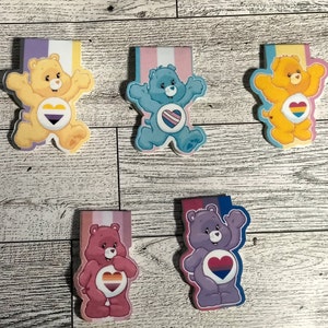 May include: Five colorful Care Bear bookmark magnets with different pride flag hearts. The magnets feature Care Bears with yellow, blue, pink, and purple fur.