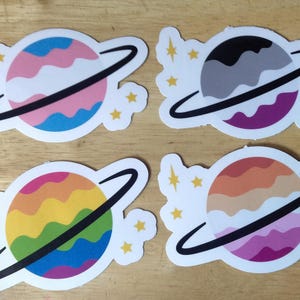 May include: Four colorful stickers featuring planets with rings. Each planet has a different color scheme, including pink, blue, gray, purple, orange, and brown. The stickers are on a wooden surface.