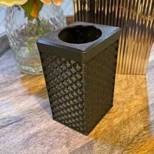 Puede incluir: Un contenedor cuadrado negro con una superficie texturizada y una abertura redonda en la parte superior. El contenedor está hecho de un material que parece ser plástico o resina.