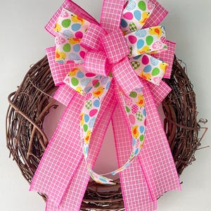 May include: A decorative Easter wreath featuring a woven grapevine base. The wreath is adorned with a large pink bow with a white grid pattern. The bow has a central ribbon with colorful Easter eggs and chicks.