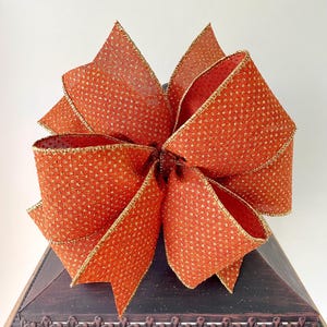May include: A large, decorative bow made of orange fabric with a gold dot pattern. The edges are trimmed with gold. The bow is likely for holiday or gift decoration.