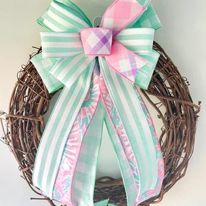 May include: A decorative grapevine wreath featuring a large bow. The bow is made of ribbons in pastel colors, including mint green, pink, and white. The ribbons have striped, plaid, and tie-dye patterns. The wreath is suitable for spring or Easter decor.
