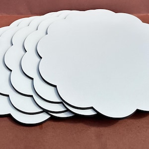 Könnte beinhalten: Ein Stapel weißer, wolkenförmiger Untersetzer mit schwarzen Rändern. Die Untersetzer sind fächerförmig angeordnet, wobei die weißen Oberflächen nach oben zeigen. Der Hintergrund ist einfarbig rotbraun.