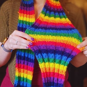 May include: A person wearing a rainbow striped knitted scarf. The scarf is made of colorful yarn and has a long, flowing design.