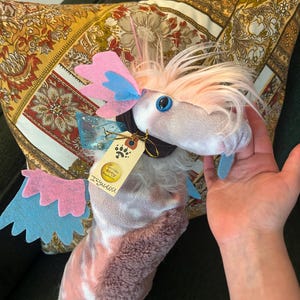 May include: A plush creature with a light pink and white patterned body, featuring soft pink hair, blue felt wings, and a blue tongue. It has a dark brown collar with a beige tag that reads "Ilywanu" and a paw print logo, plus blue plastic eyes.