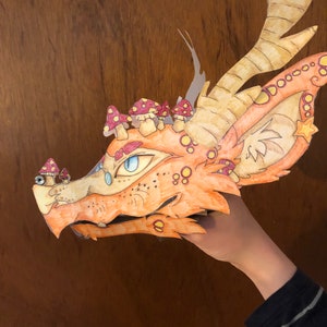 May include: A handcrafted paper dragon mask in shades of orange and yellow, featuring intricate illustrations of red and white spotted mushrooms growing along the snout and head, large pointed ears, and a single blue eye with a detailed iris.