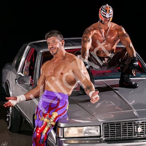 Puede incluir: Dos luchadores posan sobre un coche plateado. Un luchador lleva una máscara negra y naranja y pantalones negros. El otro luchador lleva pantalones morados y rojos con llamas.