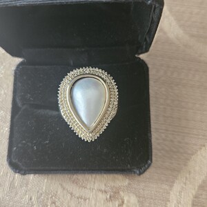 May include: A pear-shaped ring with a large, iridescent, white pearl center. The pearl is surrounded by a gold-colored frame with small, clear stones. The ring is displayed in a black velvet ring box.