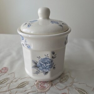 May include: A white ceramic sugar bowl with a lid, featuring a blue rose floral design. The bowl has an octagonal shape and a small knob on the lid. The floral pattern is repeated around the rim and on the lid. A decorative piece for the kitchen.