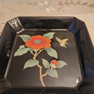 May include: A black, square decorative plate with a gold trim. The plate features a floral design with a large orange flower, green leaves, and a gold butterfly. The design is painted on the plate.