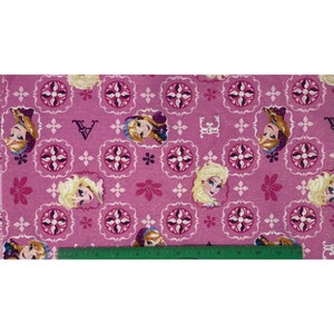 May include: Pink fabric featuring a repeating pattern of Elsa from Frozen, floral designs, and decorative elements. The fabric has a ruler along the bottom edge, with measurements up to 12 inches.