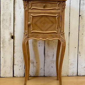 Puede incluir: Una mesita de noche de estilo antiguo, de color marrón claro, con un solo cajón y una puerta pequeña. La mesa tiene tallas ornamentadas y patas curvas. El cajón y la puerta tienen pomos de madera. La mesa mide aproximadamente 76 cm de alto.