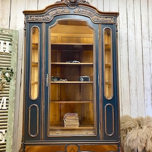 Puede incluir: Una vitrina antigua con acabado azul oscuro y marrón. El armario tiene una puerta de cristal, estantes interiores y tallas ornamentadas. El armario está lleno de cámaras y libros antiguos. El armario mide aproximadamente 183 cm de altura.