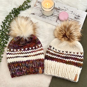 May include: Two knitted winter hats with faux fur pom poms. The hats have a white and brown striped pattern with a purple and orange color gradient. The hats have a leather tag with the word "PISH" on them.