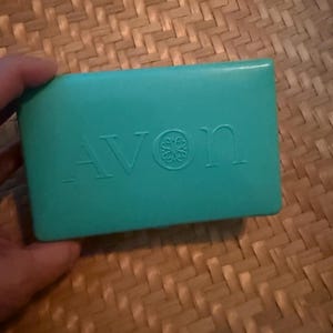 Puede incluir: Una pastilla de jabón de color turquesa con la palabra "AVON" en relieve y un diseño floral. El jabón rectangular está sostenido por una mano. El fondo es una superficie tejida de color marrón claro.
