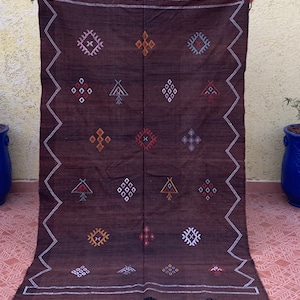 May include: A brown wool rug with a geometric pattern in shades of white, red, blue, yellow, and orange. The rug has a fringe border.