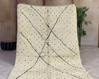 May include: A white Moroccan rug with a black diamond pattern. The rug has a fringe border and is made of wool.