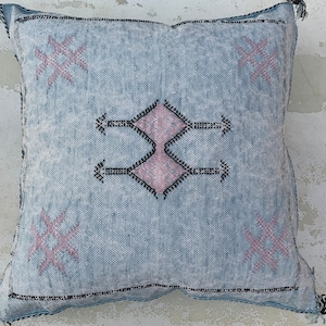 May include: A light blue throw pillow with a pink and black geometric pattern. The pillow has a fringe border.