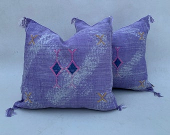 Puede incluir: Dos almohadas de tiro de color morado y blanco con borlas. Las almohadas tienen un diseño geométrico con acentos rosados y azules.