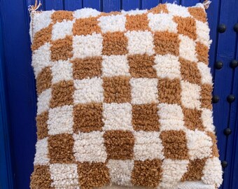 May include: A brown and white checkered throw pillow with a fluffy texture. The pillow is sitting on a woven stool in front of a blue wooden door.
