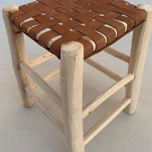 May include: A wooden stool with a woven leather seat. The stool has a simple design with four legs and two crossbars. The leather seat is brown and has a woven pattern.