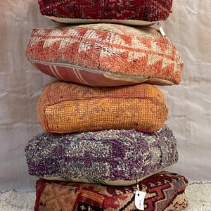 May include: A stack of five colorful Moroccan poufs with intricate patterns. The poufs are made of woven fabric and are in various shades of red, orange, purple, and green.