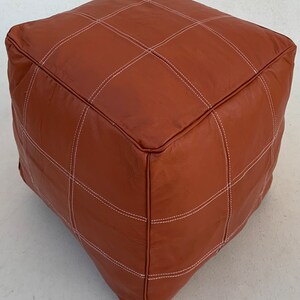 May include: A brown leather ottoman with white stitching. The ottoman has a square shape and is made of a soft, supple leather.