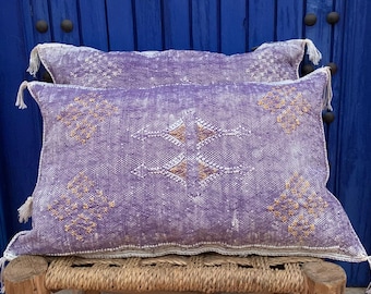 Puede incluir: Dos cojines decorativos rectangulares morados con patrones bordados en blanco y dorado. Los cojines tienen borlas blancas en las esquinas y están colocados sobre un banco tejido. El fondo es una puerta de madera azul.