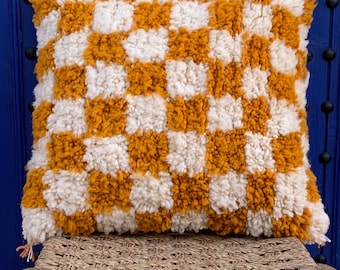 May include: A square-shaped throw pillow with a checkered pattern in mustard yellow and white. The pillow is made of a fluffy, textured fabric and is sitting on a woven wicker stool.
