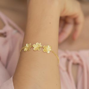 May include: A gold bracelet with three flower-shaped charms. The chain bracelet is displayed on an arm, with the flowers equally spaced. The jewellery is a simple, elegant piece.
