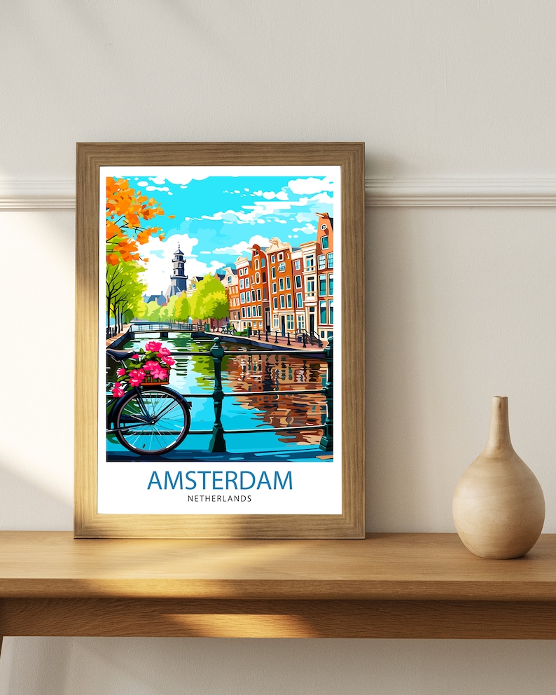 May include: A framed poster with a colorful illustration of a canal in Amsterdam, Netherlands. The poster features a bicycle with a basket of flowers, traditional buildings, and a blue sky with white clouds. The text "AMSTERDAM NETHERLANDS" is printed at the bottom of the poster.