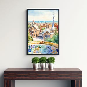 May include: A framed watercolor print of a cityscape featuring architectural details and a mosaic-tiled bench. The artwork showcases buildings with unique designs, a blue and white tower, and a view of the sea under a blue sky. The print is displayed on a dark wooden table.