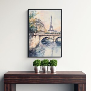 Op de afbeelding: Een aquarel schilderij van de Eiffeltoren in Parijs, Frankrijk. Het schilderij toont een brug over een rivier met gebouwen op de achtergrond. Het schilderij is ingelijst in een zwart frame en hangt aan een witte muur.