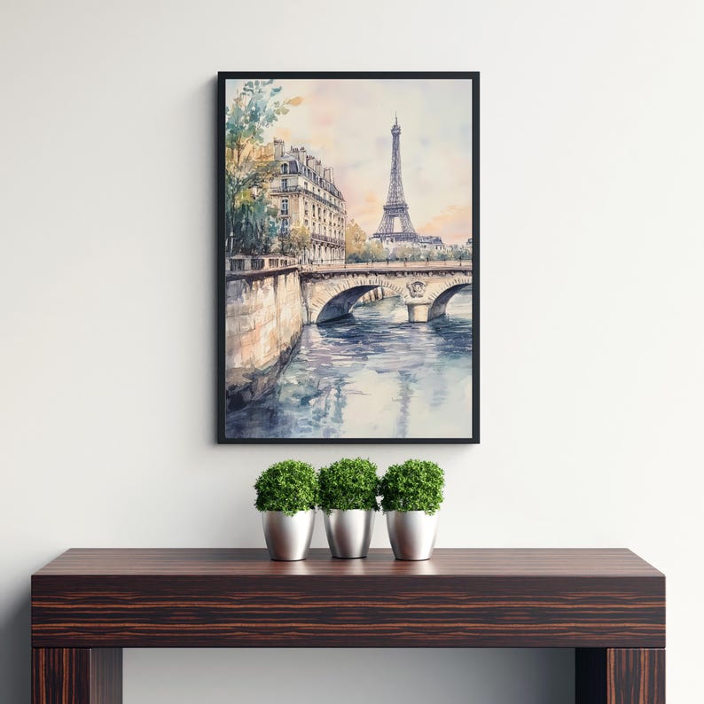 Op de afbeelding: Een aquarel schilderij van de Eiffeltoren in Parijs, Frankrijk. Het schilderij toont een brug over een rivier met gebouwen op de achtergrond. Het schilderij is ingelijst in een zwart frame en hangt aan een witte muur.