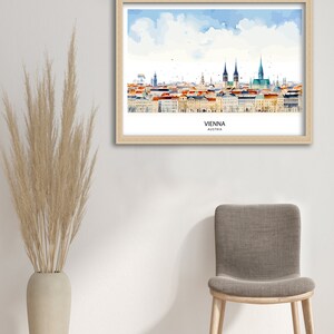Peut inclure: Une peinture &agrave; l'aquarelle de l'horizon de Vienne, encadr&eacute;e dans un cadre en bois clair. La peinture repr&eacute;sente les b&acirc;timents embl&eacute;matiques de la ville, notamment la cath&eacute;drale Saint-&Eacute;tienne et le palais Hofburg. La peinture est plac&eacute;e sur un fond de ciel bleu avec des nuages blancs. Le texte "VIENNE, AUTRICHE" est imprim&eacute; sous la peinture.