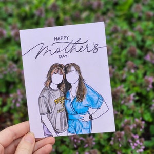 May include: A white card with the text "Happy Mother's Day" in black cursive writing. The card features a hand-drawn illustration of two women standing side-by-side. The woman on the left is wearing a gray t-shirt with the text "TACOMA" in yellow and black. The woman on the right is wearing a blue dress.