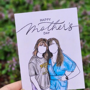 May include: A white card with the text "Happy Mother's Day" in black cursive writing. Two women are drawn on the card, one wearing a gray shirt with the text "TACOMA" in yellow and the other wearing a blue dress.