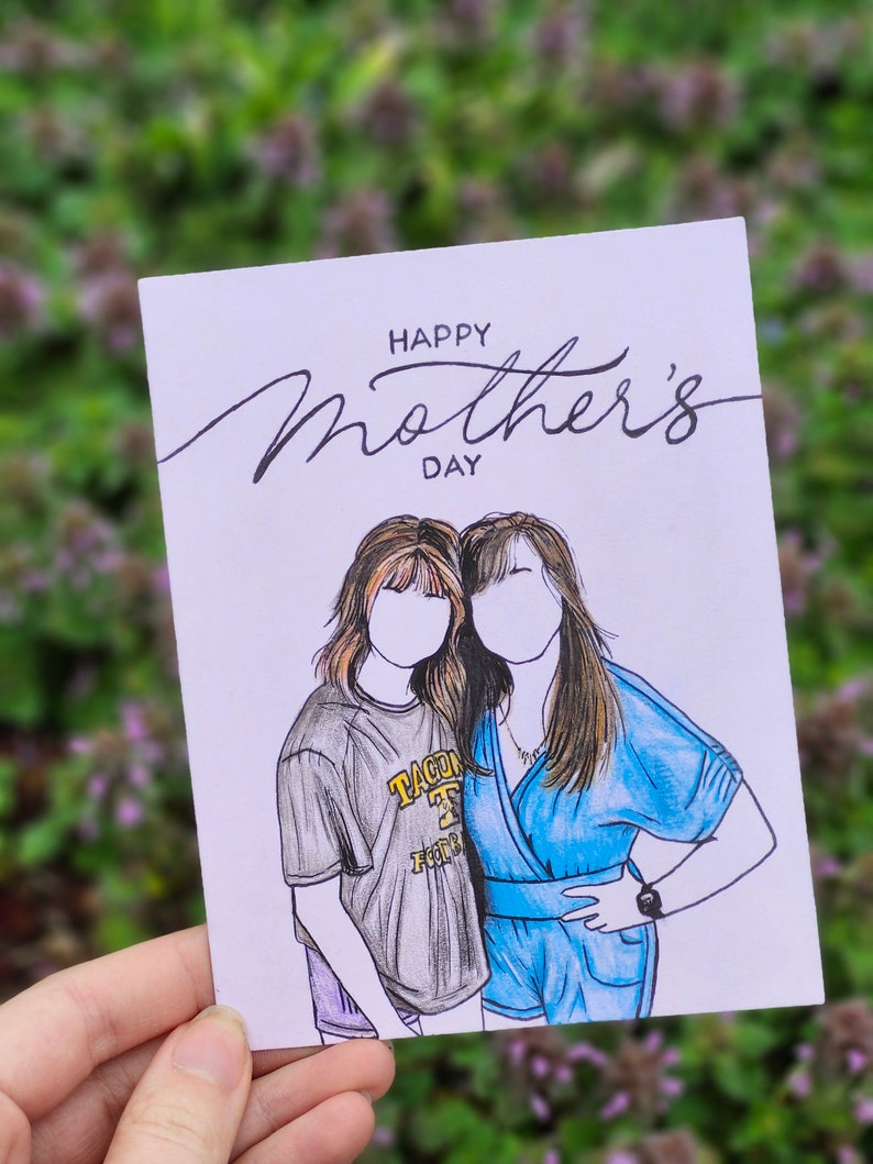 May include: A white card with the text "Happy Mother's Day" in black cursive writing. Two women are drawn on the card, one wearing a gray shirt with the text "TACOMA" in yellow and the other wearing a blue dress.